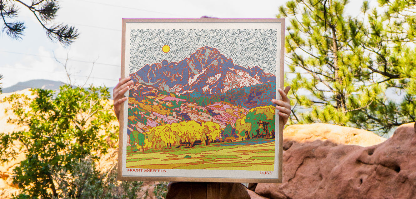 Person holding a framed artwork of a Mount Sneffles in a Colorado setting with trees and rocks.