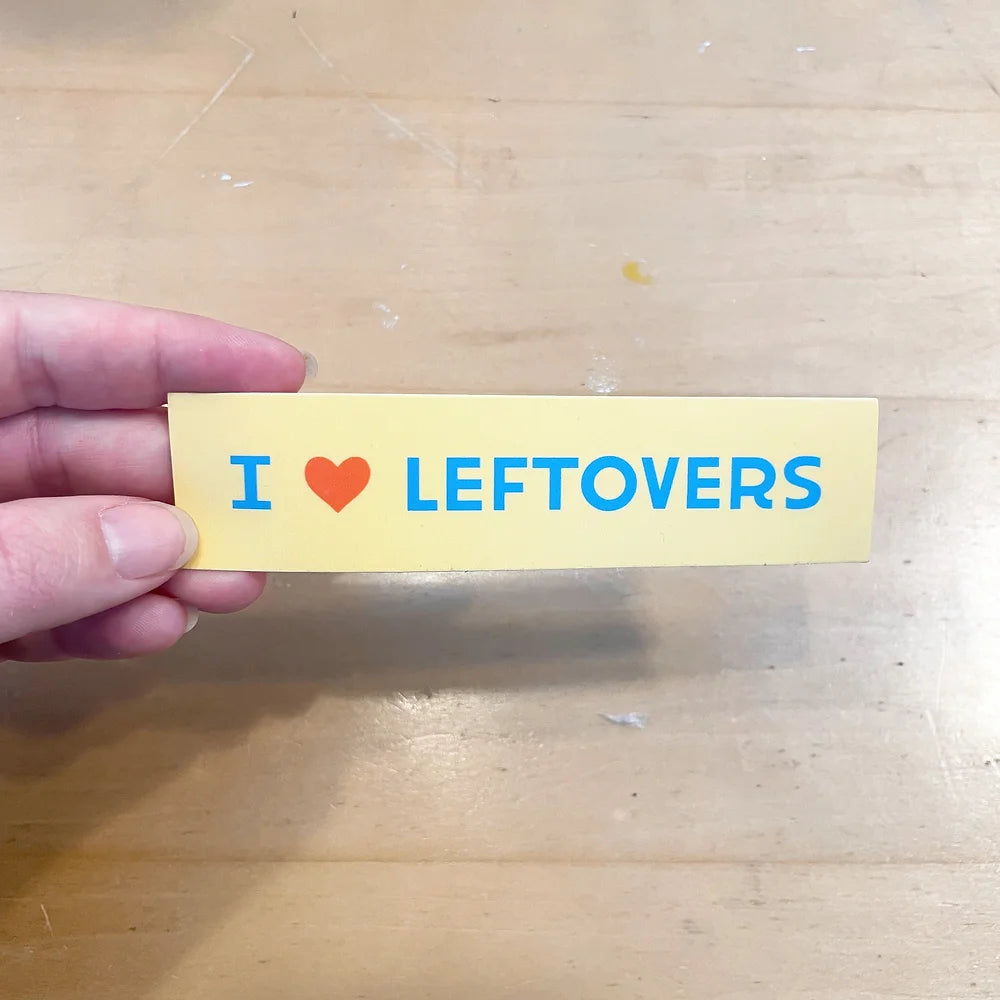 Rectangular 5” by 1.25” size yellow vinyl sticker with light blue text with a red heart that reads “I (heart) Leftovers” text. The sticker is held by somebody’s hand against a light wooden background.