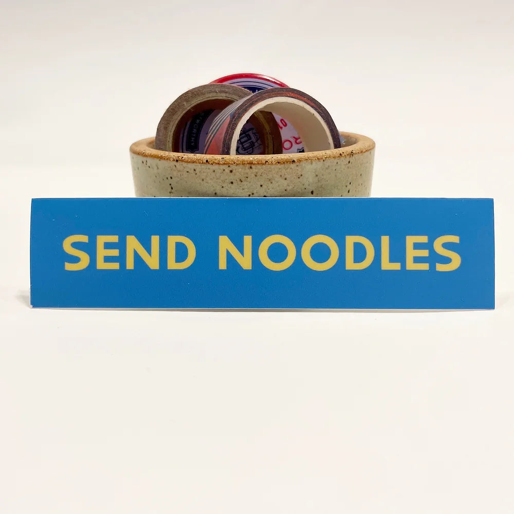 A rectangular 5” by 1.25” size vinyl blue sticker with yellow block “Send Noodles” text. The sticker is leaning against a small ceramic cup filled with washi tape and one button pin.