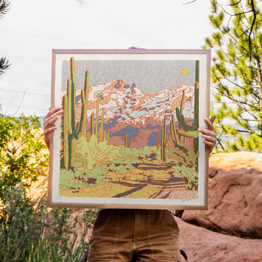 Person holding a framed artwork of a desert landscape with cacti in the Cataline desert against a natural background.