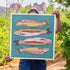 Person holding a colorful art print with trout fish illustrations in a natural setting