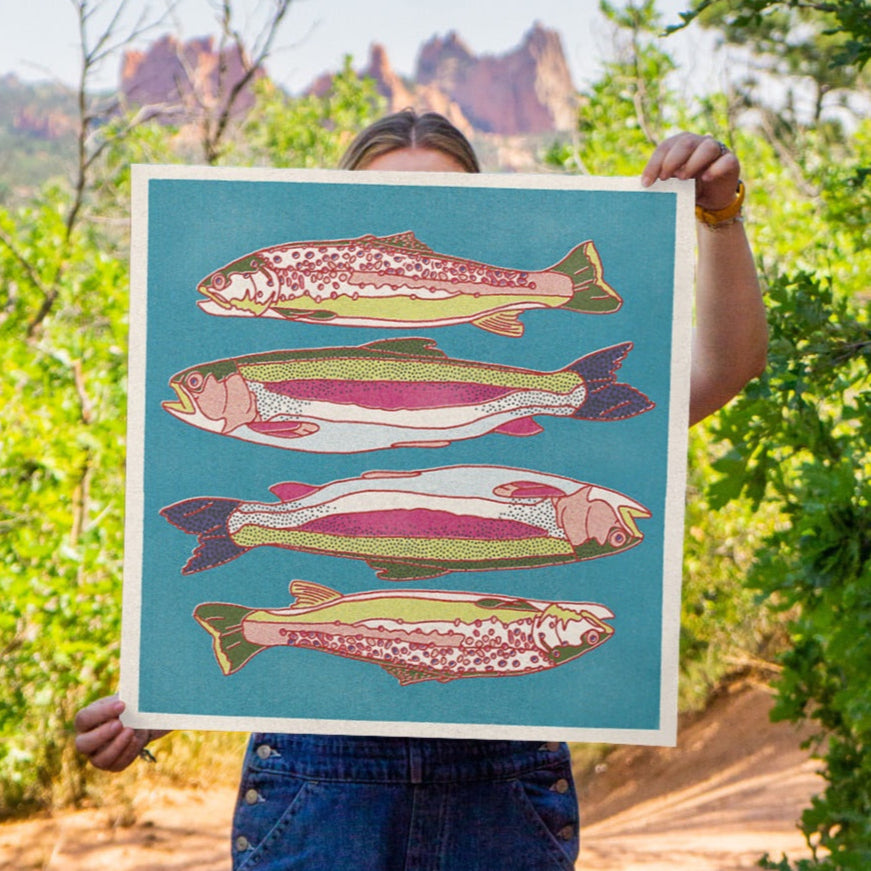Person holding a colorful art print with trout fish illustrations in a natural setting