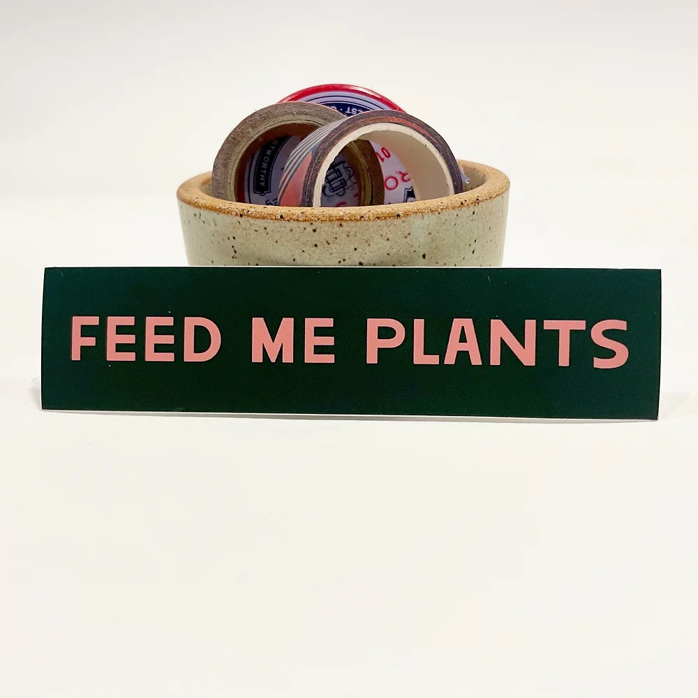 Rectangular 5” by 1.25” size vinyl black sticker with pink block “Feed Me Plants” text. The sticker is leaning against a small ceramic cup that is filled with washi tape and one button pin.
