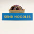 A rectangular 5” by 1.25” size vinyl blue sticker with yellow block “Send Noodles” text. The sticker is leaning against a small ceramic cup filled with washi tape and one button pin.
