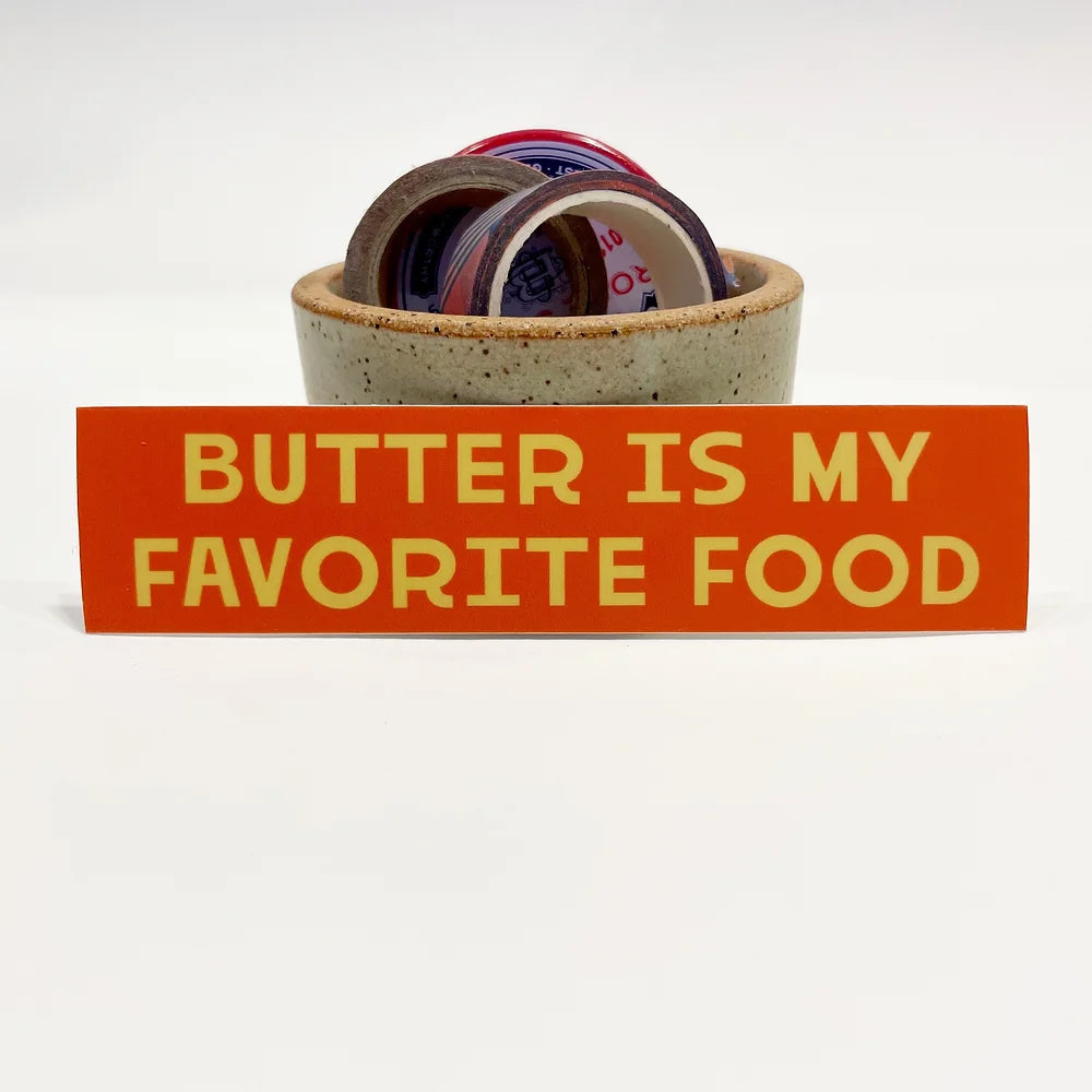 Rectangular 5” by 1.5” size red vinyl sticker with yellow block “Butter Is My Favorite Food” text. The sticker is leaning against a small ceramic cup filled with washi tape and one button pin.