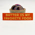 Rectangular 5” by 1.5” size red vinyl sticker with yellow block “Butter Is My Favorite Food” text. The sticker is leaning against a small ceramic cup filled with washi tape and one button pin.
