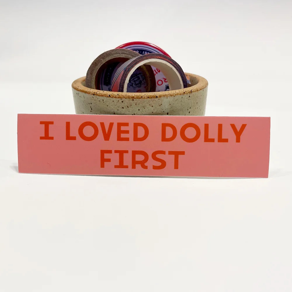 Rectangular 5” by 1.25” pink vinyl sticker with red block “I Loved Dolly First” text. The sticker is leaning against a ceramic cup filled with washi tape and a button pin.