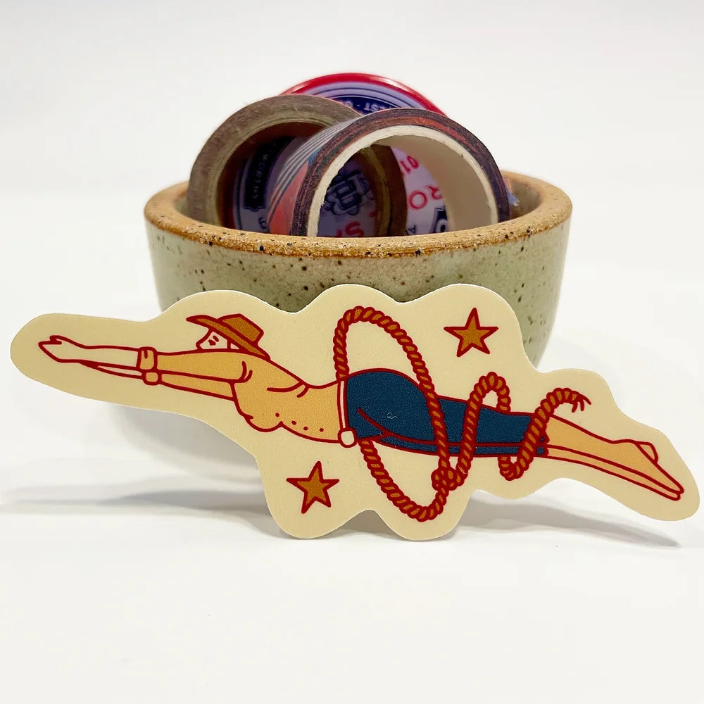 Woman diving through a red lasso. The woman is wearing a brown cowgirl hat, light yellow button up long sleeve, navy jeans, and light yellow boots that go up to her calves. There are two stars to the top right and bottom left framing the cowgirl. The background sticker color behind the cowgirl is light cream. The sticker is leaning against a small ceramic cup. The cup is filled with a variety of washi tape and a button pin.